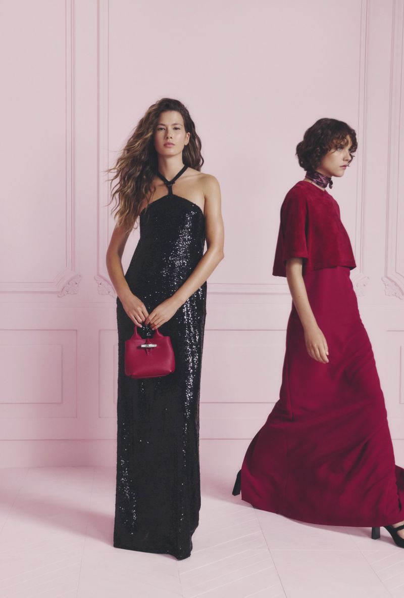 Two women pose in evening gowns against a pink background. One in a black sequin dress holds a red purse; the other wears a red gown.
