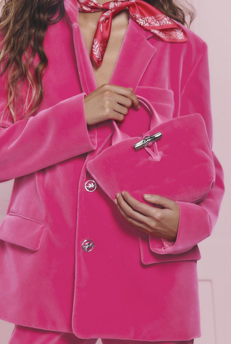 A person wears a bright pink velvet suit and a patterned scarf, holding a matching pink velvet handbag against a pink background.