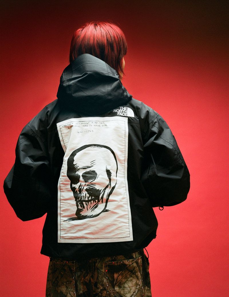 Back view of a person wearing a black hooded jacket with a skull graphic on the back, set against a solid red backdrop.