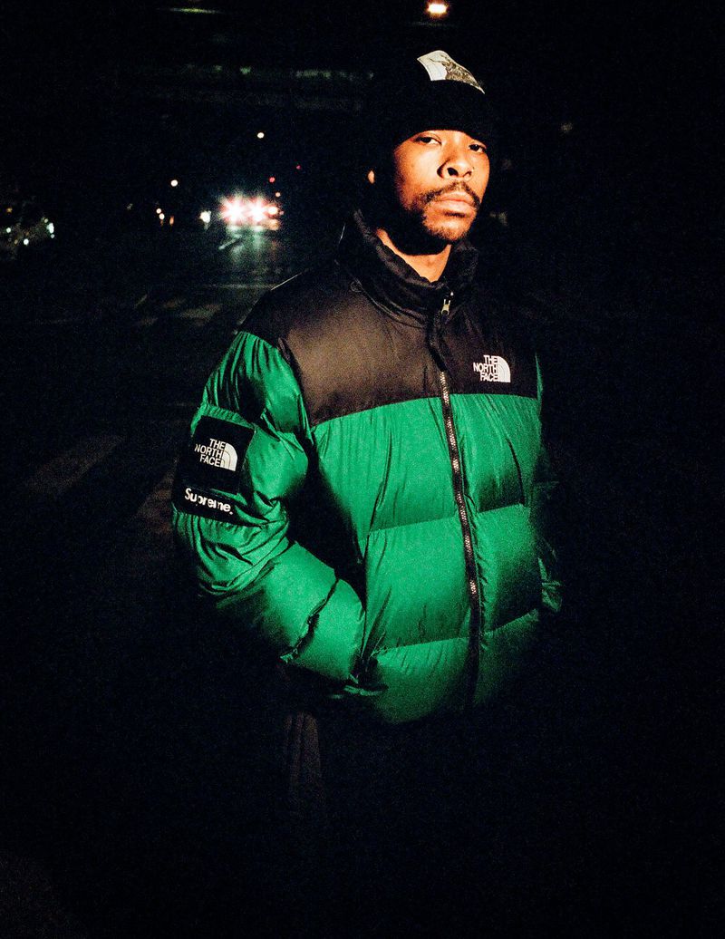 A man stands outdoors in a green and black puffer jacket and black beanie against a dark background at night with car headlights.