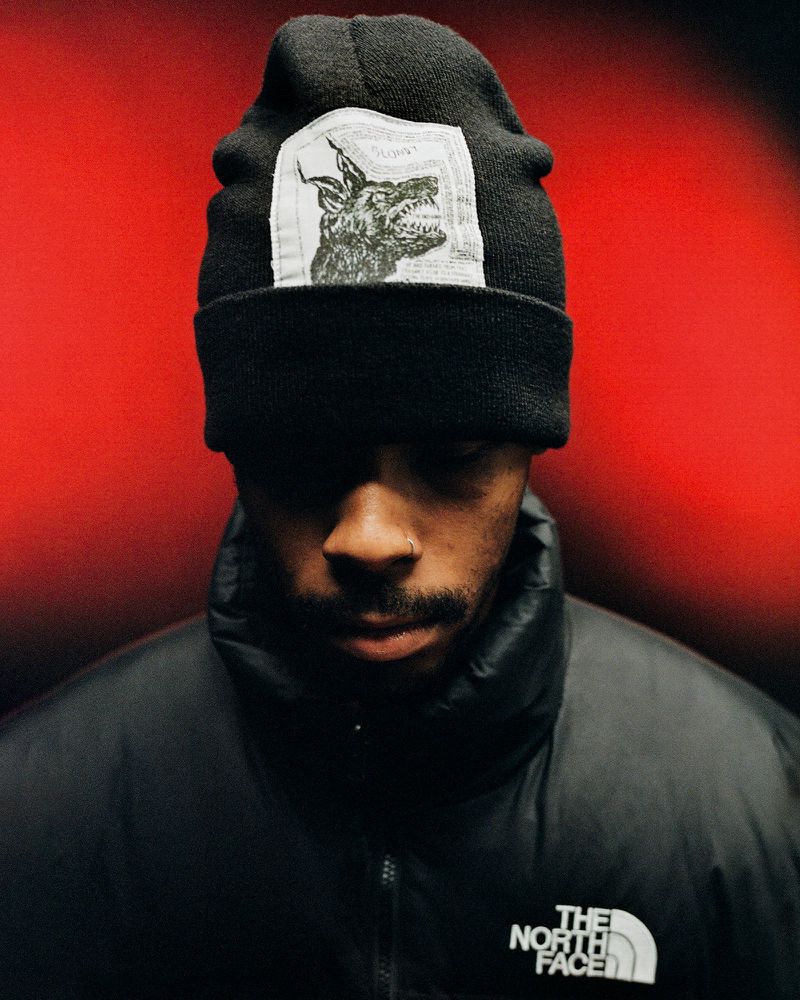 Close-up portrait of a young adult wearing a black beanie with a wolf graphic and a black puffer jacket against a red backdrop.
