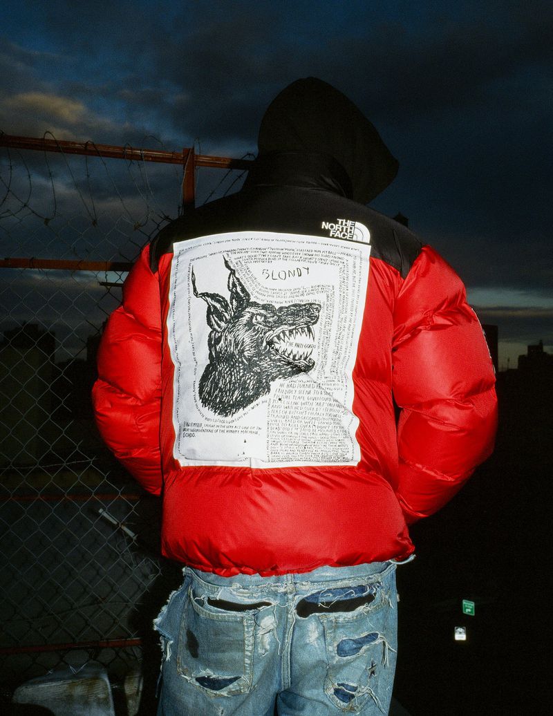 Person wearing a red and black North Face puffer jacket with a dog graphic, distressed jeans, standing outdoors at dusk. 