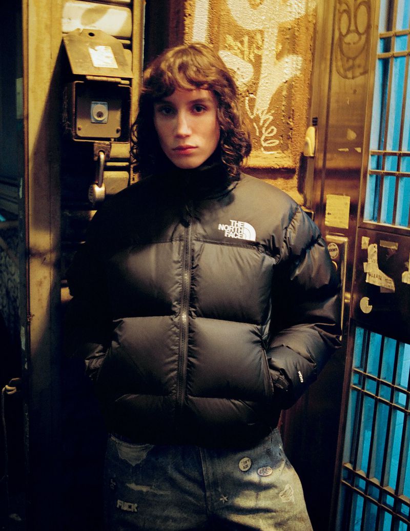 Portrait of a young adult with shoulder-length brown hair wearing a black puffer jacket and patterned jeans against an urban backdrop.