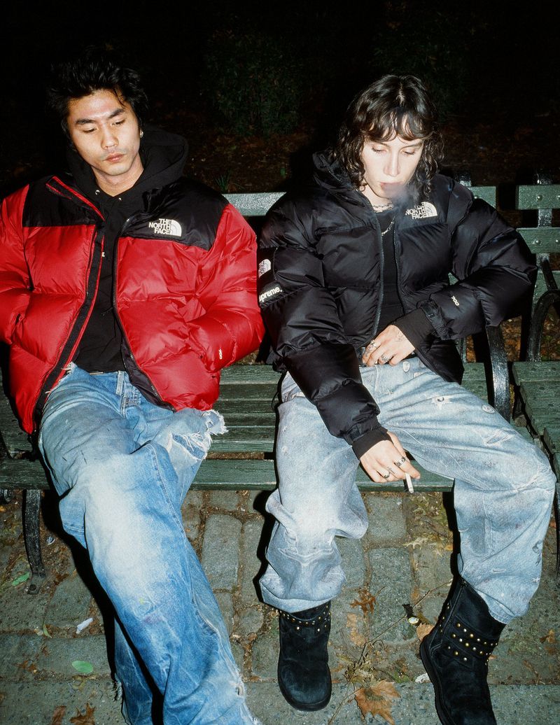 Two people in puffer jackets sit on a park bench at night. One is smoking. Both wear jeans and boots in an urban outdoor scene.