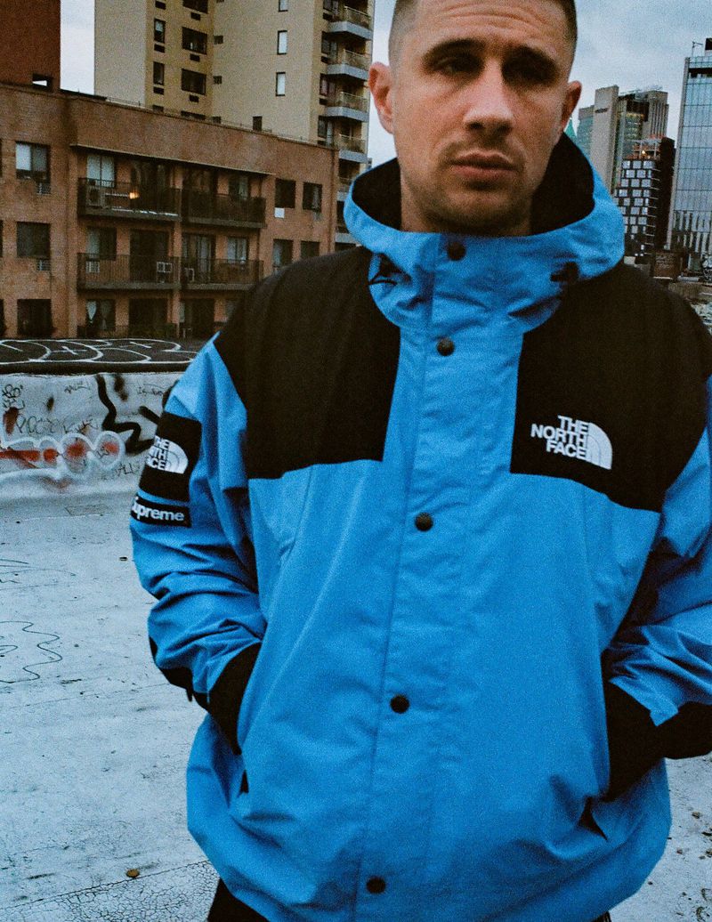 Man in blue and black North Face jacket outdoors on urban rooftop with city buildings in the background. Fashion and lifestyle photo.