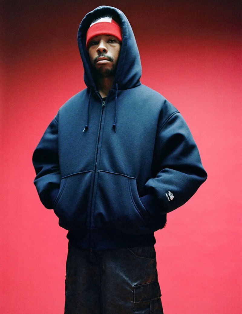 Man in a blue hooded zip-up sweatshirt and dark pants poses against a red background, wearing a red headband, looking at camera.
