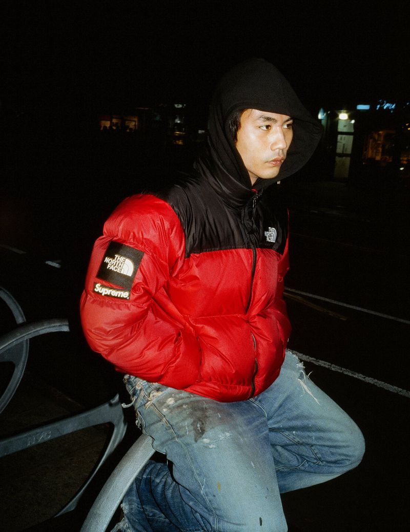 A young Asian man poses in a red and black North Face puffer jacket at night, sitting on a metal bar in an urban environment.