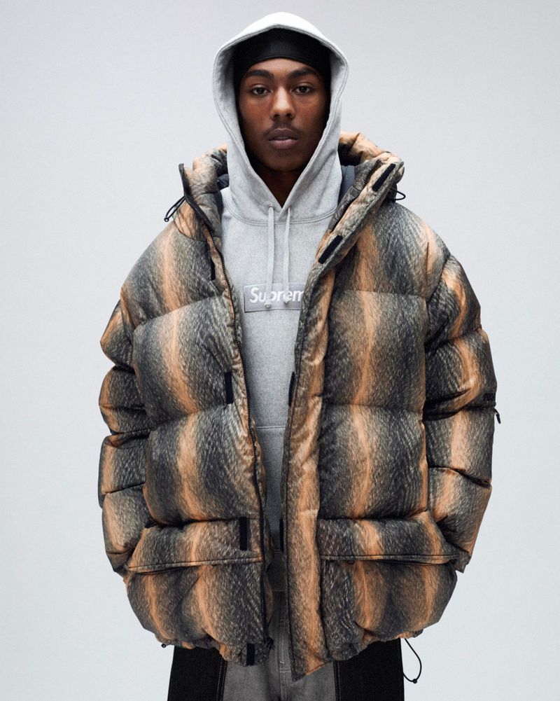 Person wearing a hooded gray sweatshirt and a large puffer jacket with brown and gray stripes standing against a white background.