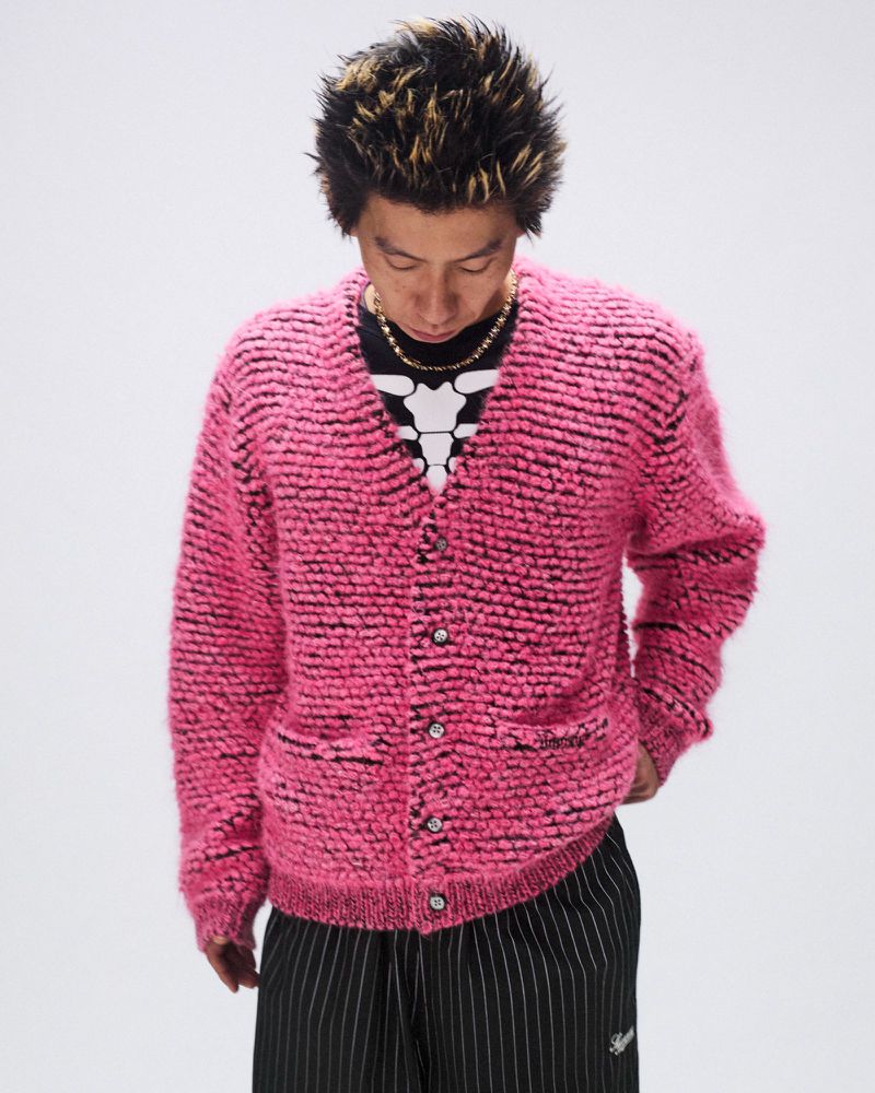 Adult male in a pink textured cardigan, black and white striped pants, and gold chain necklace in a studio setting.