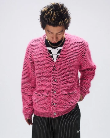 Adult male in a pink textured cardigan, black and white striped pants, and gold chain necklace in a studio setting.