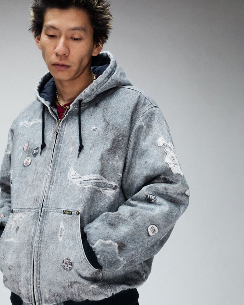 Young adult in hooded denim jacket with pins, posing indoors with a neutral expression, in front of a plain light-gray backdrop.