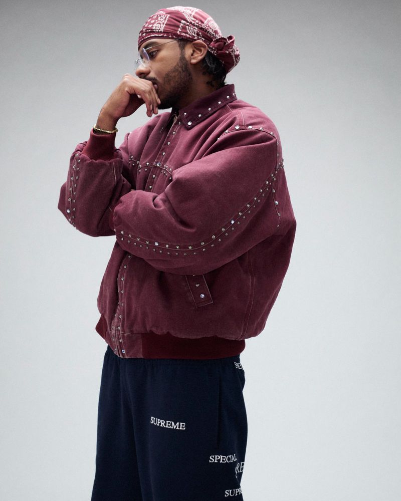 Adult man in a maroon jacket with silver embellishments poses indoors, wearing dark sweatpants and a maroon patterned headscarf.