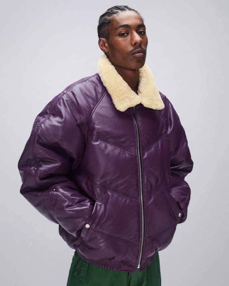 Man in purple puffer jacket with fur collar poses in a studio setting. Fashionable winter outerwear with a zipper and button details.