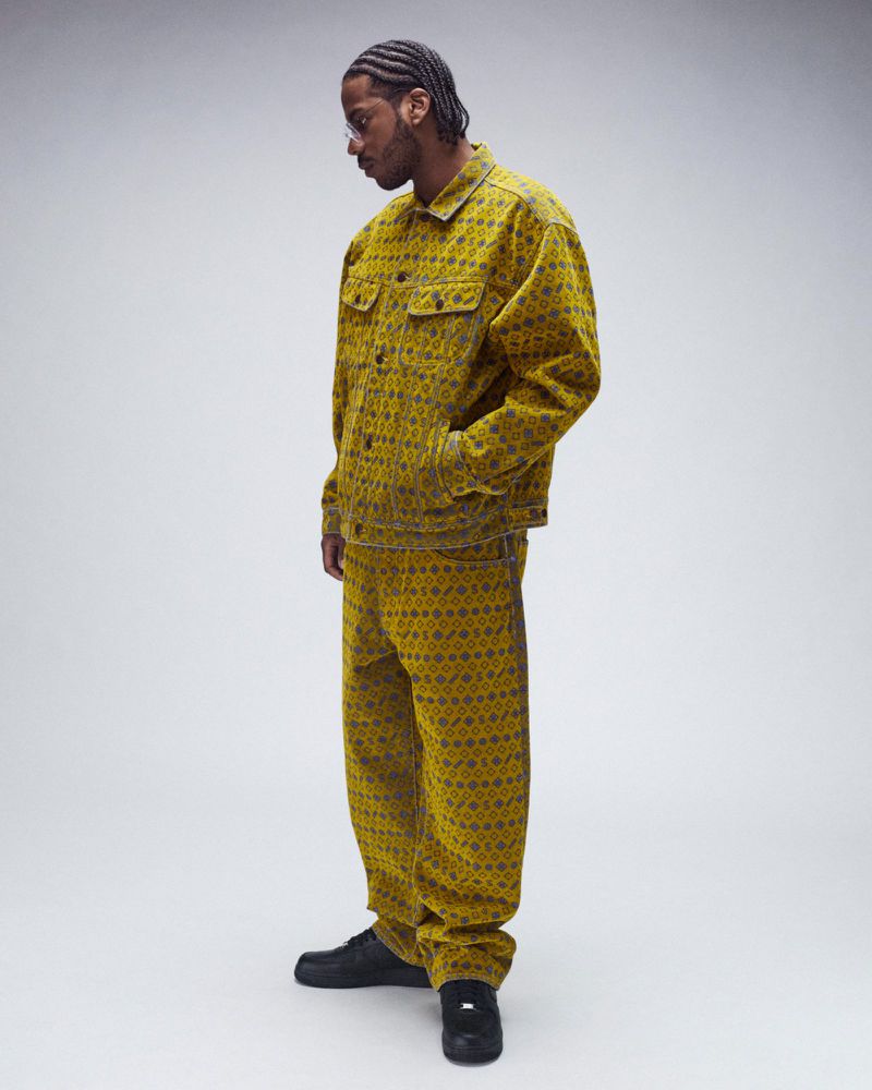 Man in patterned denim jacket and pants, standing indoors. He is looking down with a neutral expression in front of a grey background.
