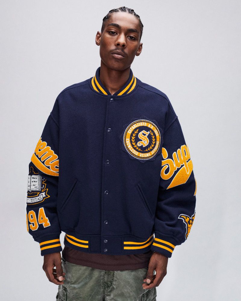 Adult male in a navy blue varsity jacket with yellow trim and patches, posing in a studio setting, showcasing streetwear fashion.
