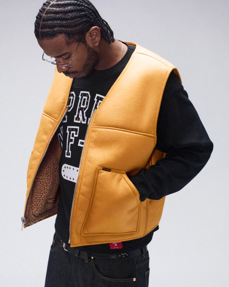 Man poses in a studio wearing a yellow fleece vest, black long-sleeve shirt, jeans, and glasses against a neutral background.