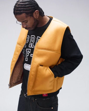 Man poses in a studio wearing a yellow fleece vest, black long-sleeve shirt, jeans, and glasses against a neutral background.