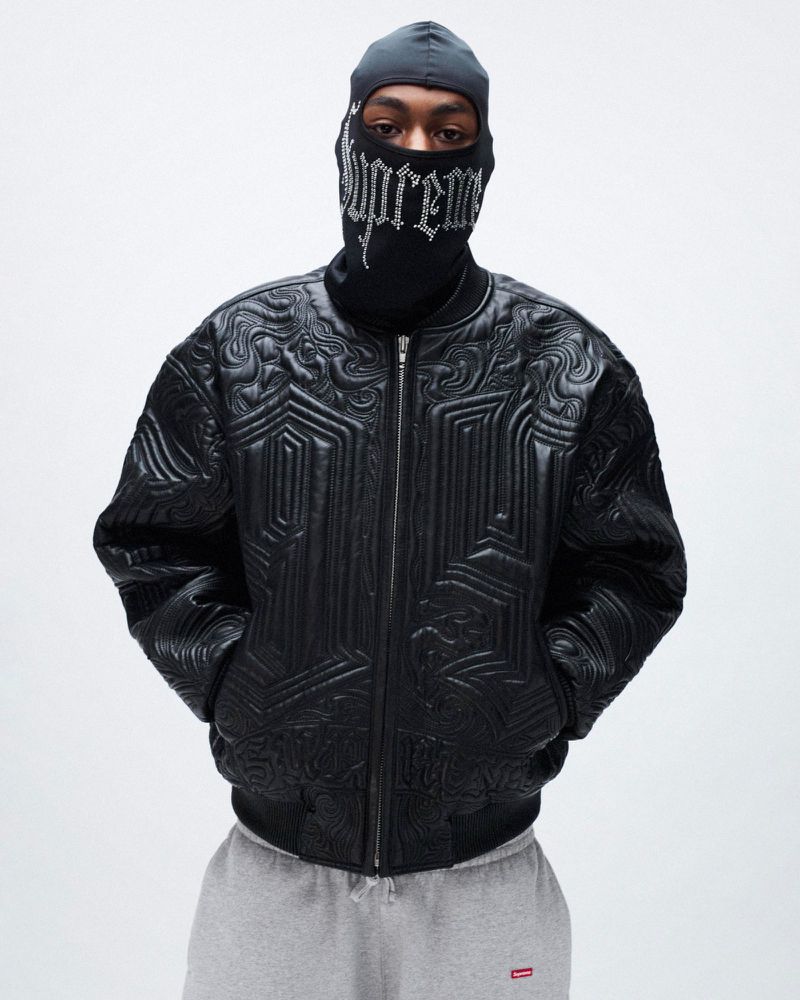 Person in balaclava and black quilted jacket, posing against white backdrop, gray sweatpants, product shot, indoor studio.