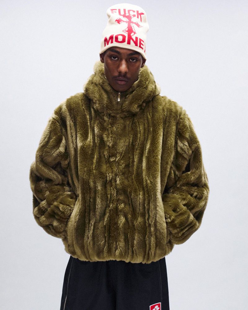 Adult male wearing a faux fur jacket and a knit cap, standing indoors against a plain white backdrop.