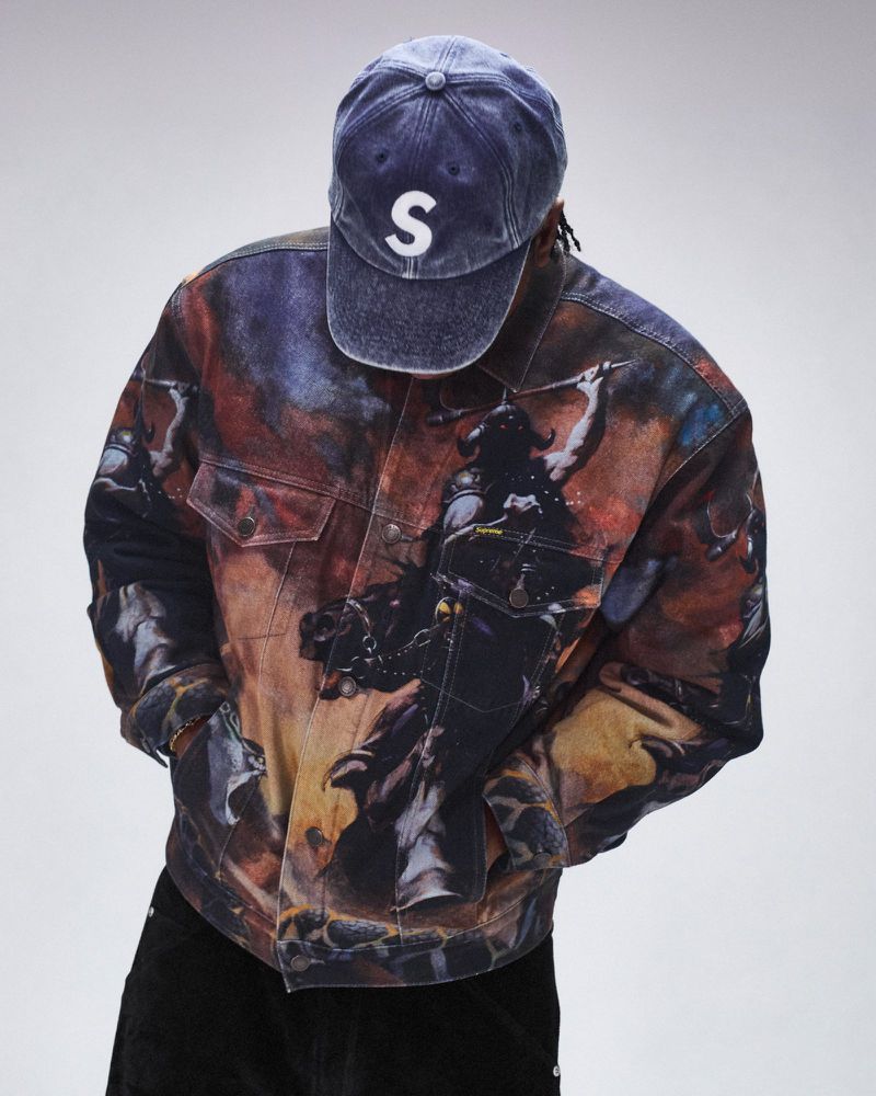 Person wearing a painterly jean jacket with a blue cap, set against a solid white backdrop in a studio-style fashion shot.