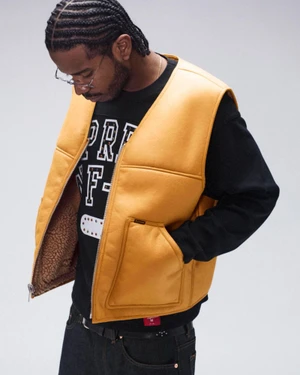 Black man wearing a yellow vest over a black graphic sweatshirt and dark jeans against a neutral white background