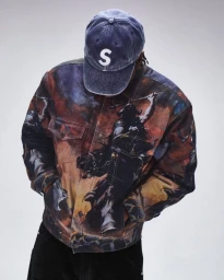 Model wearing a printed jacket and blue washed cap with an S logo against a plain grey studio background.