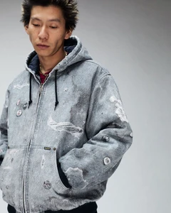 Man wearing a distressed grey zip-up hoodie with pins and patches against a neutral grey studio background