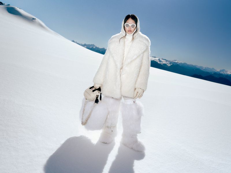 Woman in white coat and furry boots stands in snowy alpine landscape. Bright light and clear blue sky over snow-capped mountains.