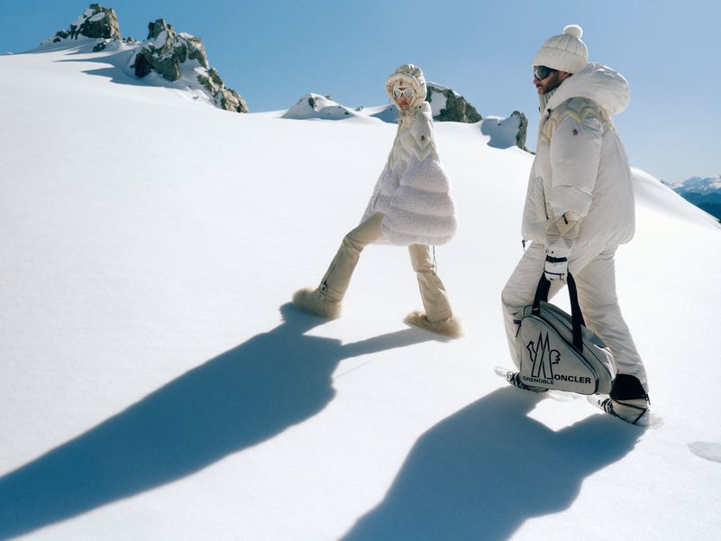 Two people in white winter apparel walk on snow-covered mountainside with blue sky; one carries a duffel bag. Fashion editorial.