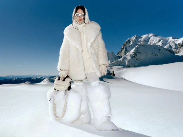 Woman in winter fashion posing in a snowy mountain landscape with white fur coat, furry leg warmers, headscarf, sunglasses and small white bag.