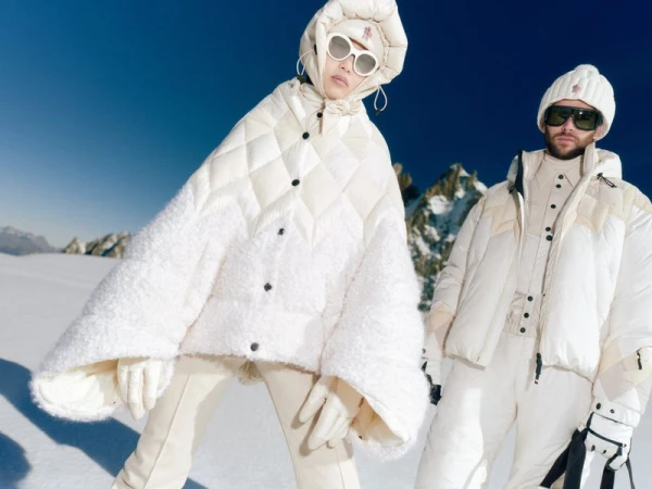 Two people in winter fashion on a snow-covered mountain, wearing white ski suits, sunglasses, and beanies, under a clear blue sky.