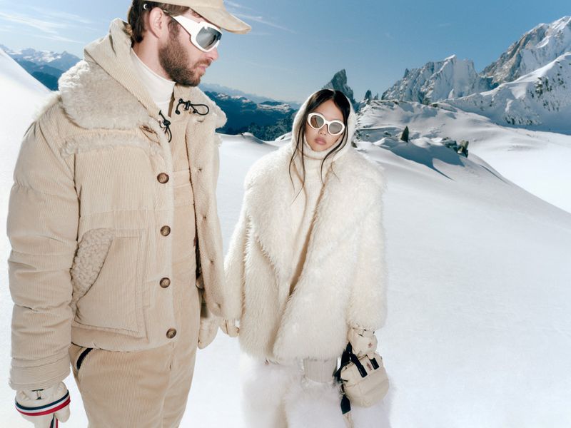 Couple in winter attire holding hands in a snowy mountain landscape. Man in corduroy, woman in fur coat and sunglasses, bright daylight.