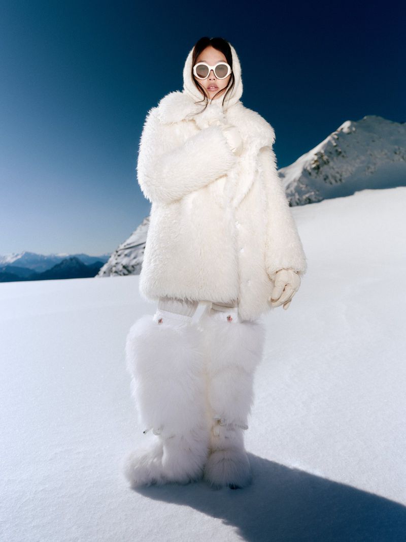 A woman in a white fur coat and sunglasses stands in a snowy landscape with mountains in the background on a bright, sunny day.