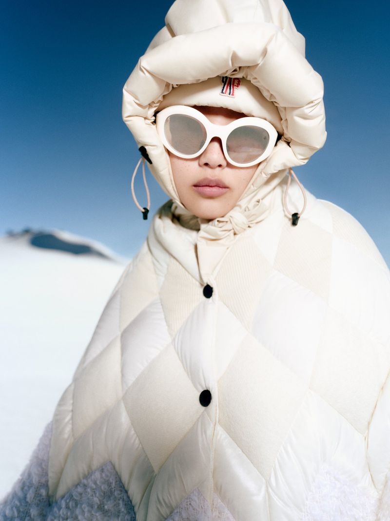 Woman in a quilted off-white hooded jacket and sunglasses stands out against a bright blue sky and snowy landscape in a fashion shot.