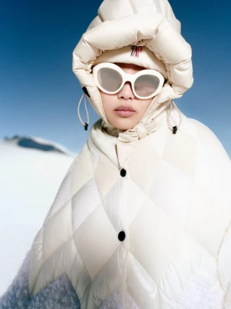 Woman in a quilted off-white hooded jacket and sunglasses stands out against a bright blue sky and snowy landscape in a fashion shot.