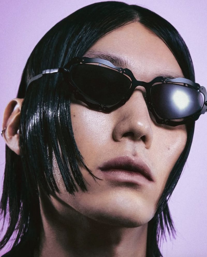 Portrait of a person wearing black sunglasses and with shiny black hair, in front of a light purple background.