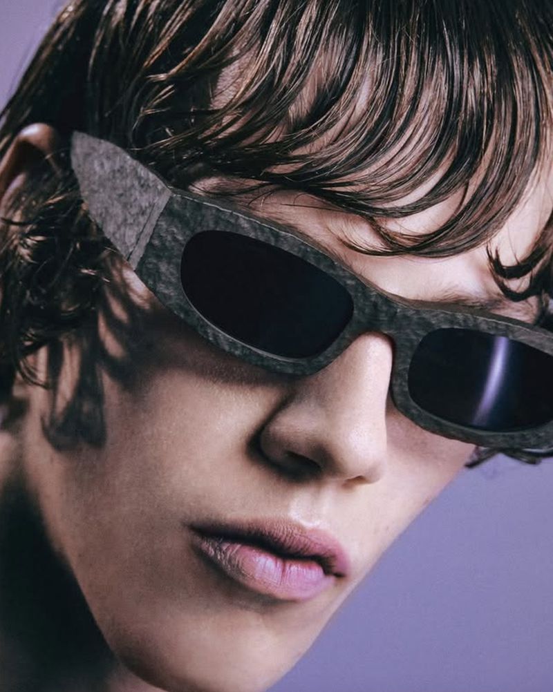 Close-up of an adult with brown wavy hair wearing gray sunglasses with black lenses against a light purple backdrop.