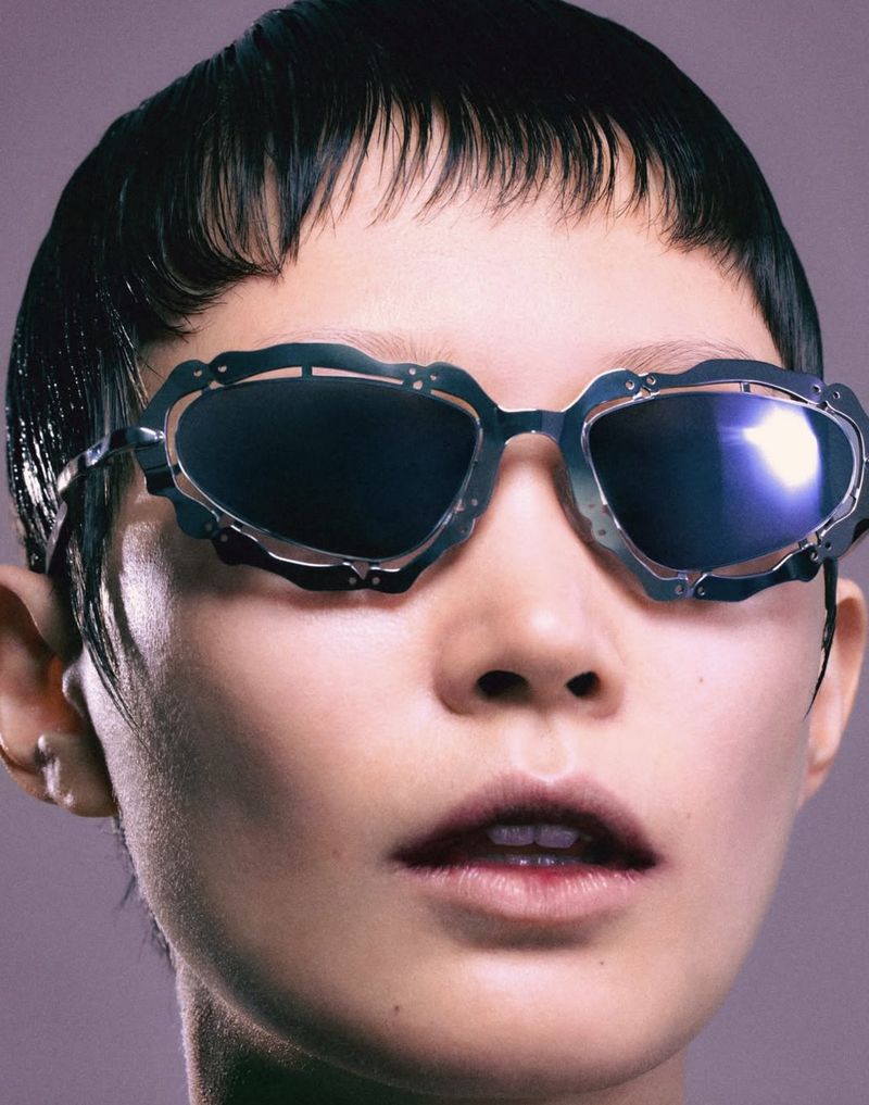 Close-up of a fair-skinned person with black hair wearing sunglasses with metallic frames on a neutral gray background.