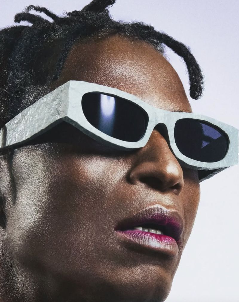 Portrait of adult woman with dark skin and dreadlocks wearing oversized, geometric sunglasses against a gradient purple backdrop.