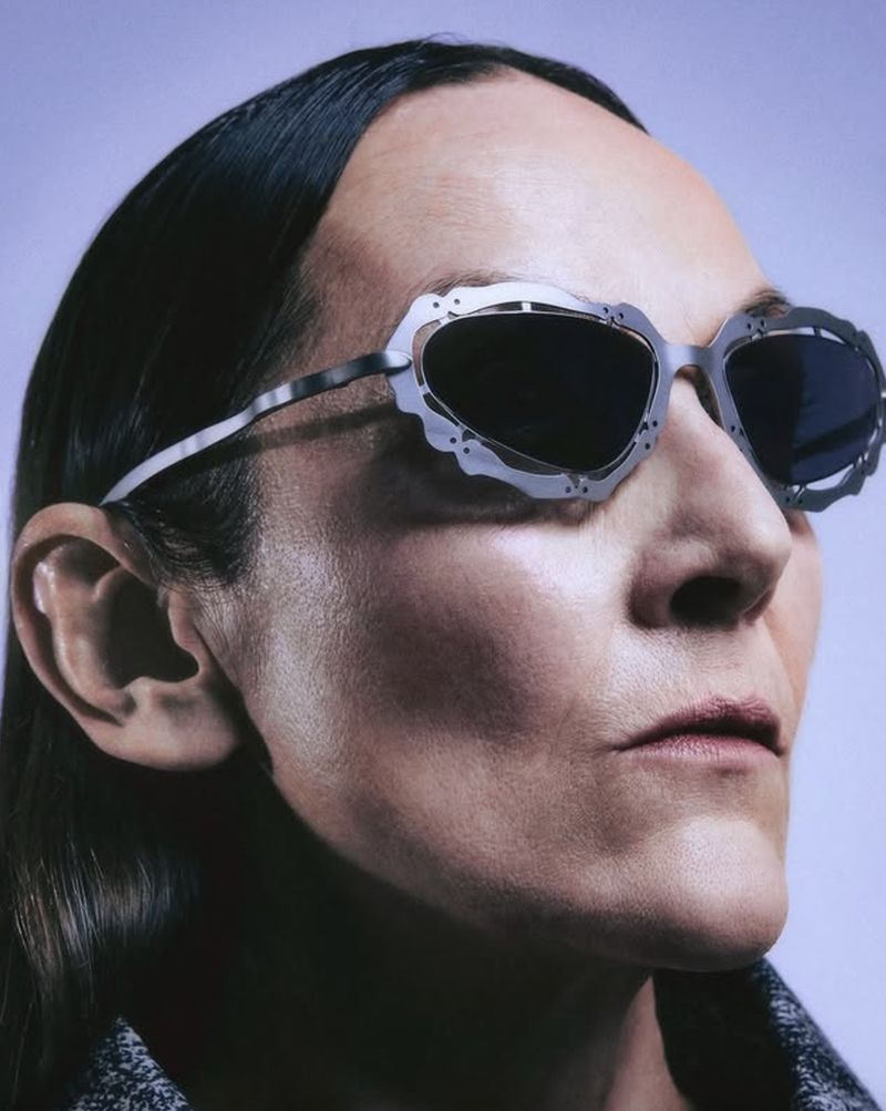 Close-up of a person wearing avant-garde sunglasses with a light gray metal frame and dark lenses against a light background.