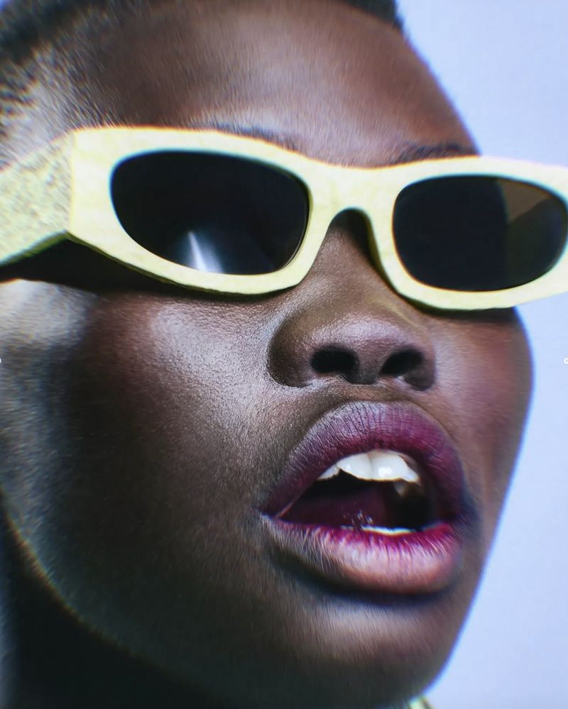 Close-up shot of a dark-skinned adult female model wearing fashionable, lemon-yellow sunglasses and deep purple lipstick, expressing with an open mouth.