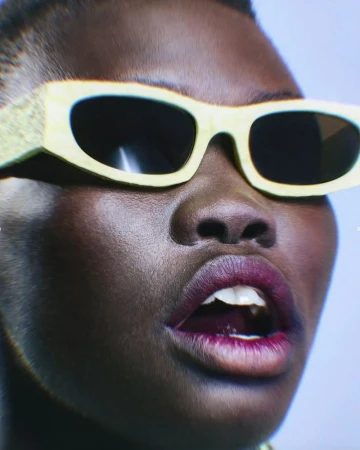 Close-up shot of a dark-skinned adult female model wearing fashionable, lemon-yellow sunglasses and deep purple lipstick, expressing with an open mouth.