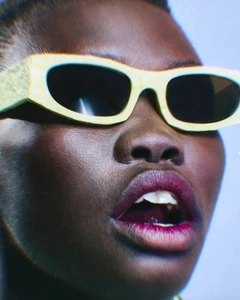 Close-up portrait of a model wearing bold yellow sunglasses and dark tinted lenses against a plain pale background