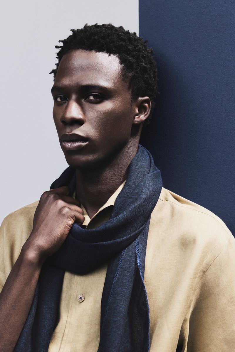 Black male model in a beige button-up shirt and dark navy scarf against a split blue and gray backdrop.