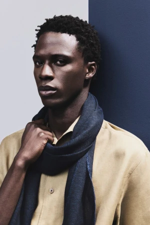 Black male model in a beige button-up shirt and dark navy scarf against a split blue and gray backdrop.