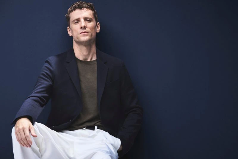 Portrait of adult man in dark blazer and white pants, casually seated against a navy blue backdrop, looking at the camera.