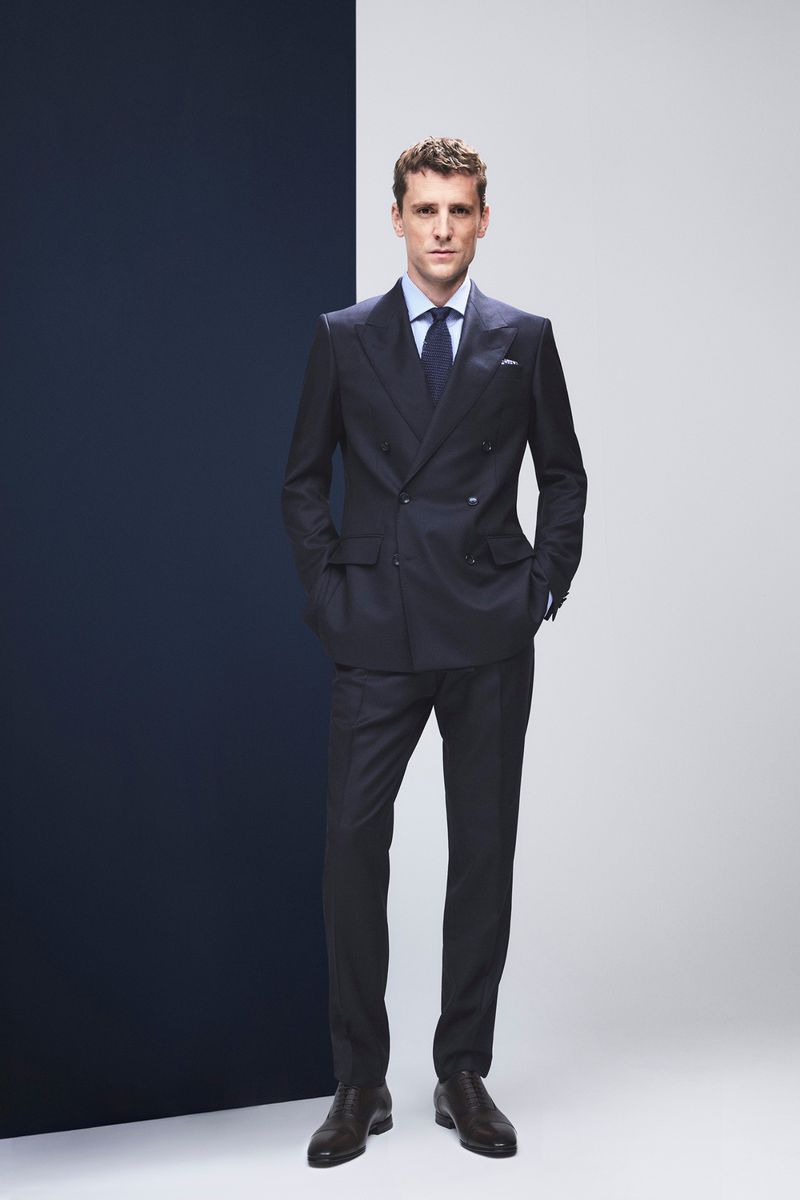 Full-length shot of a man in a dark suit and tie, standing in front of a vertically divided background of white and dark blue.
