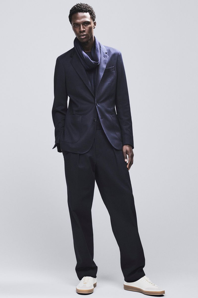 A stylish African American man in a navy suit and scarf poses against a light grey backdrop in a fashion editorial-style full shot.