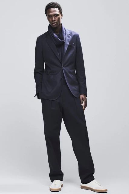 A stylish African American man in a navy suit and scarf poses against a light grey backdrop in a fashion editorial-style full shot.