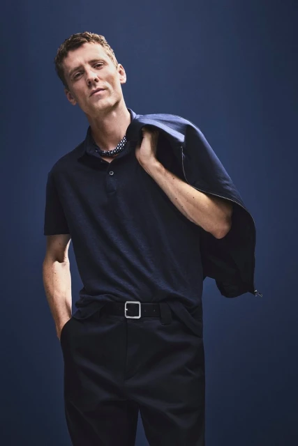 Man in navy polo shirt with jacket slung over shoulder, poses against dark blue backdrop in eye-level indoor portrait.
